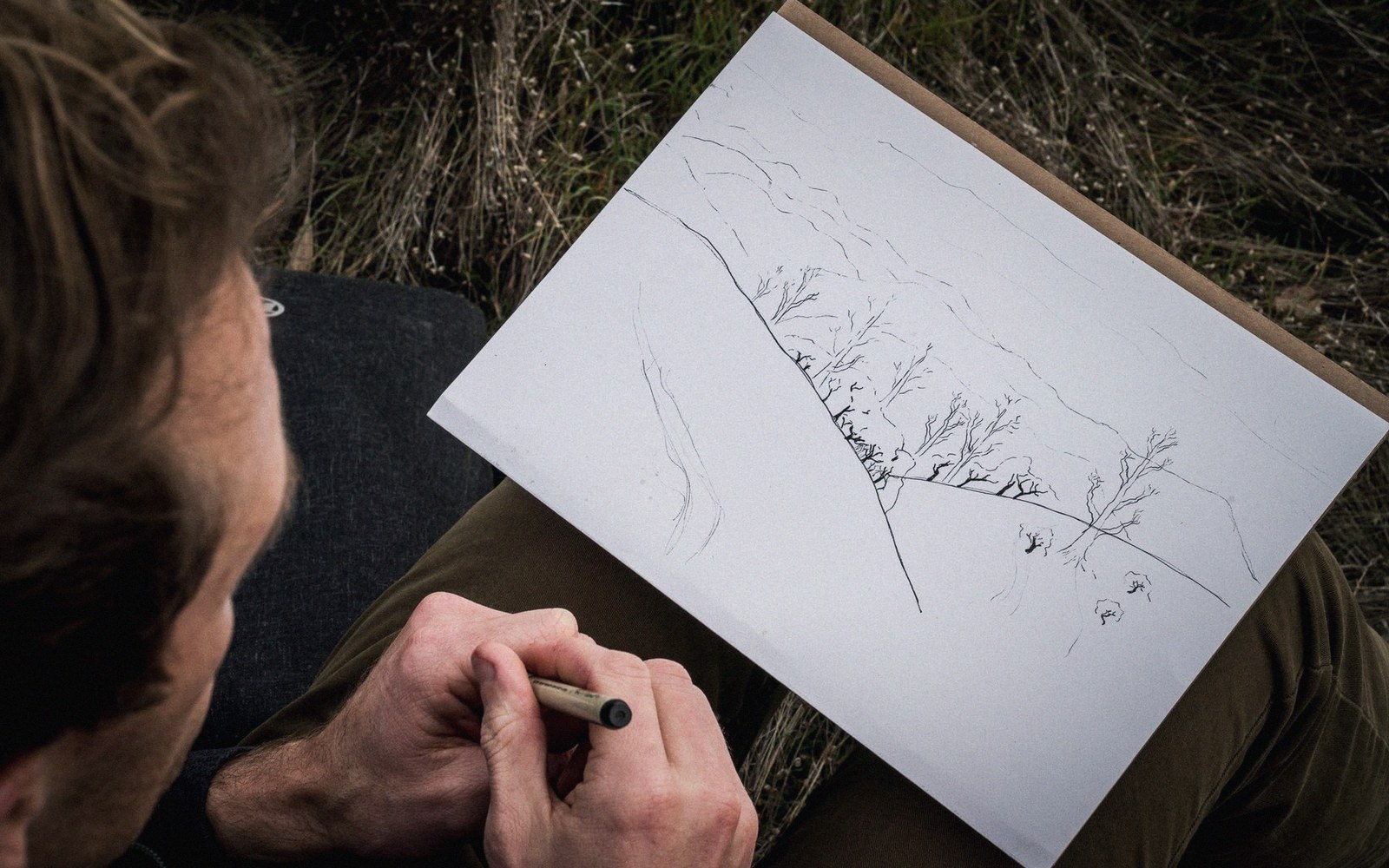 A person sketches a landscape with bare trees on a pad while sitting outdoors